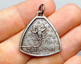 May include: Silver-toned pendant with a triangular shape, featuring a scorpion design surrounded by stars. The pendant includes the dates 24.10 and 22.11, and the astrological symbol for Scorpio. The pendant has a loop at the top for a chain.