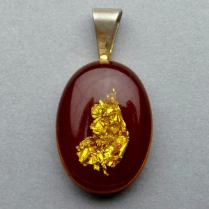 Puede incluir: Colgante ovalado con una fianza plateada y un centro rojo oscuro. El centro presenta un grupo de pan de oro, creando un contraste llamativo. El colgante es una joya, adecuado para collares u otros accesorios.