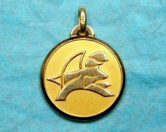 May include: A gold-colored pendant featuring a stylized image of an archer with a bow and arrow. The pendant is round and has a smooth, polished surface.