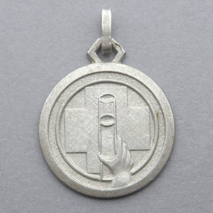 May include: Silver pendant featuring a red cross and a hand holding a medical syringe. The pendant is circular and has a raised edge.