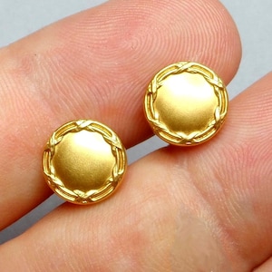 Könnte beinhalten: Zwei goldfarbene, runde Knöpfe. Jeder Knopf hat einen zentralen, flachen Kreis, der von einem geflochtenen, seilartigen Rand umgeben ist. Die Knöpfe werden vor einem neutralen Hintergrund gezeigt.