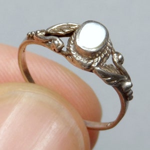 Può includere: Un anello d'argento con una pietra ovale bianca incastonata in un design delicato e ornato. L'anello presenta dettagli intricati, tra cui foglie e volute.
