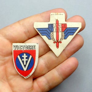 May include: Two vintage paper pins with patriotic designs. One pin is shield-shaped with the word "VICTOIRE" and a cross symbol. The other pin is arrow-shaped with the words "FRANCE LIBRE" and a sword design. Both pins have red, white, and blue colors.