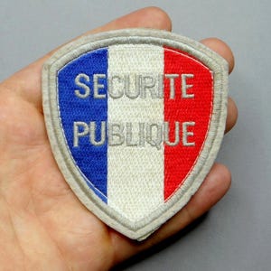 Peut inclure: Un écusson brodé blanc, bleu et rouge avec le texte "SECURITE PUBLIQUE" en lettres argentées. L'écusson est en forme de bouclier et a un fond blanc avec une bande bleue à gauche et une bande rouge à droite.
