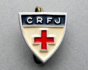 Puede incluir: Un broche vintage en forma de escudo con un emblema de cruz roja. El broche presenta un centro color crema con una cruz roja, una sección superior azul marino con las letras "CRFJ" y un borde plateado. El broche tiene un pequeño lazo dorado en la parte superior.