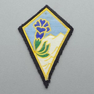 Peut inclure: Un écusson brodé en forme de losange, avec un motif de fleur stylisé en bleu, vert et jaune.