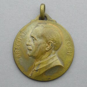May include: A gold-colored medal with a profile of a man on the front. The medal has the text "MARÉCHAL FOCH" around the edge.