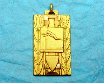 May include: Gold-colored metal pendant with a design of a person jumping over a hurdle. The pendant has a decorative border with leaves and a shield shape at the bottom.
