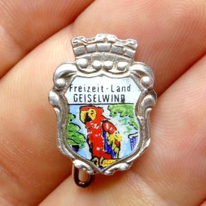 Peut inclure: Une épingle argentée avec un motif de bouclier. Le bouclier présente une illustration colorée de perroquet et le texte "Freizeit-Land GEISELWIND". L'épingle a un sommet en forme de couronne et une petite boucle en bas.