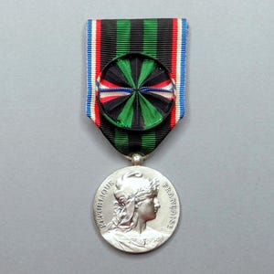 May include: A silver medal with a green, black, and white ribbon. The medal features a profile of a woman with a laurel wreath on her head. The inscription on the medal reads "REPUBLIQUE FRANCAISE".