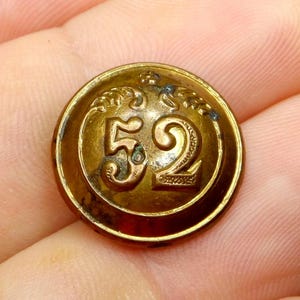 Può includere: Un bottone vintage in ottone con il numero "52" in evidenza. Il bottone circolare presenta dettagli decorativi sul bordo e intorno ai numeri. Il bottone ha un aspetto leggermente invecchiato.