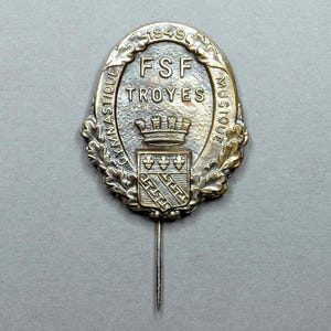 May include: A silver and gold pin with the text "FSF TROYES" and "GYMNAStique MUSIQUE" around the edge. The pin also features a crown and a coat of arms with three fleurs-de-lis. The year "1949" is inscribed at the top.