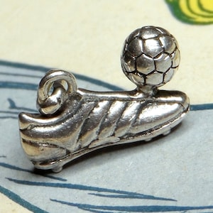 Peut inclure: Breloque en argent représentant une chaussure de football avec un ballon de football sur le dessus. Le breloque a une boucle pour l'attacher à un bracelet ou un collier.