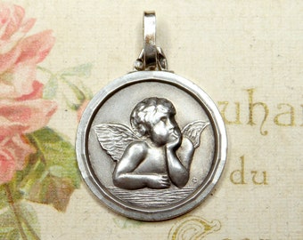 May include: A silver pendant with a cherub relief, resting its chin on its hand, with wings. The round pendant has a simple border and a loop for a chain. The background is a vintage floral design.