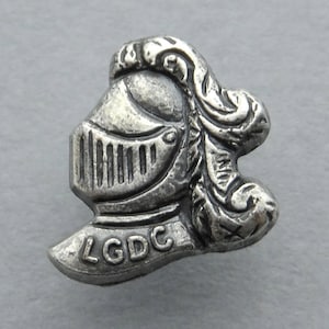 Peut inclure: Épinglette en métal argenté représentant un casque de chevalier avec des détails ornés et l'inscription "LGDC".