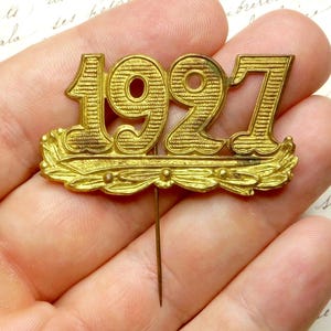 May include: A gold-toned metal pin with the year 1927 embossed on it. The pin has a decorative leaf design on either side of the year.
