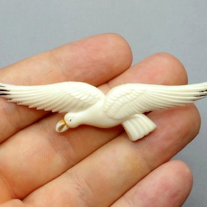 Peut inclure: Figurine d'oiseau crème aux ailes déployées, avec des détails de plumes sculptées et des accents noirs sur les extrémités des ailes. Le bec de l'oiseau est jaune clair. Il s'agit probablement d'une broche ou d'un pendentif.