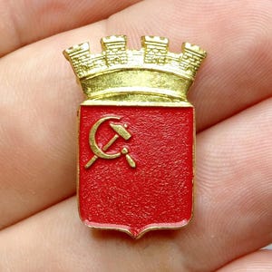 Puede incluir: Pin metálico dorado con un escudo rojo con un emblema dorado de martillo y hoz. El pin tiene una corona dorada en la parte superior. Probablemente un artículo vintage o de colección.