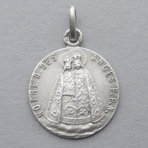 Puede incluir: Un colgante de plata con una imagen religiosa de Nuestra Señora de los Ángeles. El colgante es redondo y tiene un bucle en la parte superior para colgar. La imagen es de una mujer con una larga túnica y una corona en la cabeza. El texto "NOTRE D. DES ANGES P.P.N." está inscrito alrededor del borde del colgante.