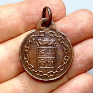 Peut inclure: Une médaille de couleur cuivre avec une bordure en chaîne et un blason au centre. Le blason présente un écu avec un oiseau, une croix et un motif à damiers.