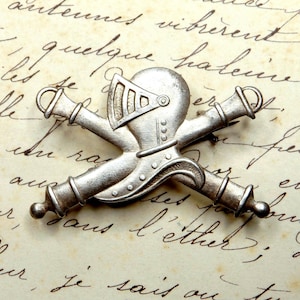 May include: A silver-toned metal pin in the shape of crossed cannons with a helmet on top. The helmet has a visor and a decorative design. The pin is on a piece of paper with handwritten text.