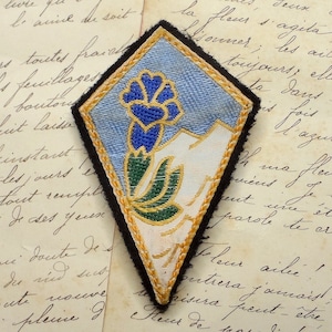 May include: A diamond-shaped patch with a blue, green, and gold embroidered flower design. The patch has a black border and a white background.