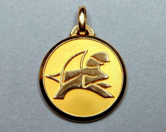 May include: A gold pendant with a stylized image of a person riding a horse. The pendant is round and has a smooth, polished surface.