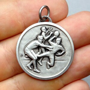 Peut inclure: Pendentif en argent avec un design tridimensionnel de trois personnages jouant au basket-ball. Le pendentif est circulaire et possède une boucle en haut pour l'attacher à une chaîne.