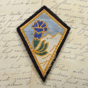 May include: A blue, green, and gold embroidered patch in the shape of a diamond. The patch features a stylized flower with a blue center and green leaves. The background is a light blue fabric with a gold border.