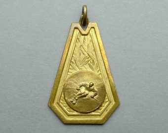 May include: A gold-colored metal medal with a skier in relief on the front. The medal is shaped like a triangle with rounded corners and has a decorative border.