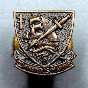 Puede incluir: Una insignia de los Comandos de la Marina de color bronce. La insignia tiene forma de escudo y presenta un barco, una espada y una cruz. Las palabras "COMMANDOS MARINE" están inscritas en la parte inferior de la insignia.