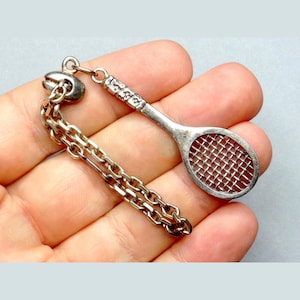 Peut inclure: Un charme de raquette de tennis en argent avec une chaîne attachée. Le charme est détaillé avec un filet en maille et un manche texturé.