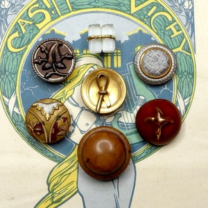 Op de afbeelding: Assortiment van vintage knopen in verschillende kleuren en ontwerpen, waaronder een gouden knoop met een gestileerd ankh-symbool, een bruine knoop met een bloemmotief, een rode knoop met een gouden kruis, een gouden knoop met een geometrisch patroon en een bruine knoop met een wit en gouden ontwerp.