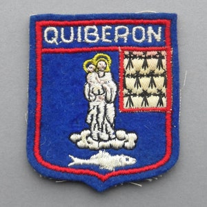 May include: A blue felt patch with red embroidered trim, featuring the text "QUIBERON" in white embroidery. The patch depicts a white embroidered image of a saint holding a baby, a white embroidered fish, and a white embroidered grid pattern.