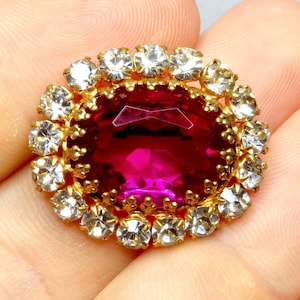 May include: A gold brooch with a large, oval, magenta gemstone at its centre, encircled by clear, round crystals. The crystals are held in place by individual gold prongs, giving a sparkling appearance. The brooch is likely vintage or antique.