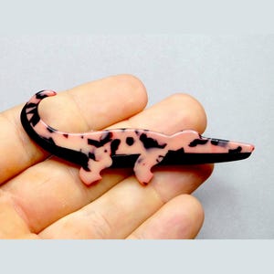Peut inclure: Broche en forme d'alligator rose et noir. L'alligator a un corps rose avec des taches noires et un ventre noir. Un accessoire unique.