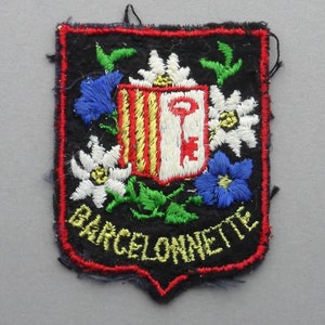 May include: A black embroidered patch with a red border. The patch features a white shield with a red and yellow striped design and a key. The shield is surrounded by white and blue flowers. The text "BARCELONNETTE" is embroidered in yellow on the bottom of the patch.