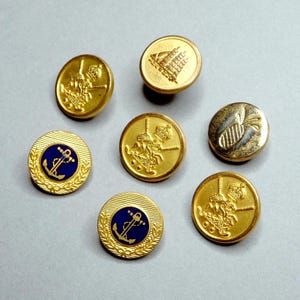 May include: A collection of gold-colored buttons with various embossed designs. Some buttons feature a knight on horseback, others a building, and some have a blue center with a gold anchor. The buttons are round and have a metallic sheen.