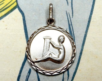 May include: A silver-colored pendant featuring the Virgo zodiac symbol. The round pendant has a textured edge and a loop for a chain. The Virgo figure is depicted holding a sheaf of wheat.