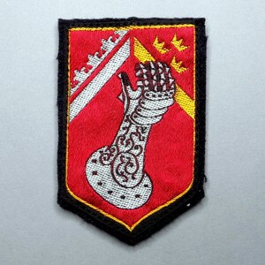 May include: A red, white, and gold embroidered patch with a black border. The patch features a white arm with a gold cuff and a gold crown on the upper arm. The arm is holding a gold object. There are five gold stars in the upper right corner of the patch.