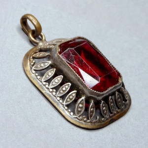 Op de afbeelding: Een vintage hanger met een grote, rechthoekige, gefacetteerde rode edelsteen. De edelsteen is gezet in een zilverkleurig metalen frame met een decoratieve goudkleurige rand en een lus voor een ketting.
