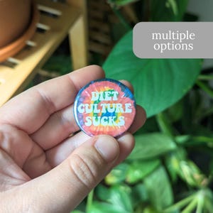 May include: A round button with the text "DIET CULTURE SUCKS" in white, set against a tie-dye background of red, orange, yellow, blue, and green. The button is held in a hand, with a blurred green plant in the background.
