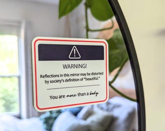 May include: A rectangular sticker with a white background, a red border, and a dark blue header featuring a white triangle warning icon. The text reads: WARNING! Reflections in this mirror may be distorted by society's definition of beautiful. You are more than a body.