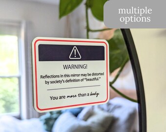 May include: A white sticker with a red border and a warning sign. The sticker reads "WARNING! Reflections in this mirror may be distorted by society's definition of "beautiful." You are more than a body."