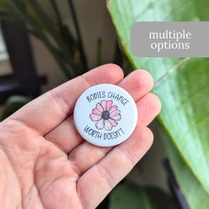 May include: White button with a pink flower and the text "Bodies Change Worth Doesn't"