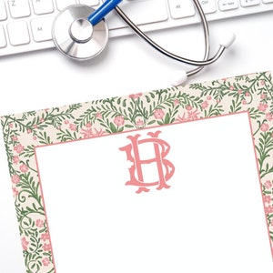 May include: A floral-bordered notepad with a pink monogram, featuring a cream background with green leaves and pink flowers. A blue stethoscope rests above the notepad, next to a white keyboard.