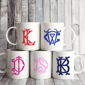 May include: Five white ceramic mugs with different colored initial monograms. The mugs are arranged in a row, with the monogrammed mugs in the center and the plain mugs on either end. The monograms are in red, blue, pink, and blue.
