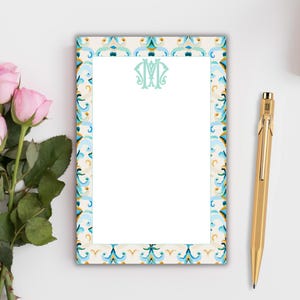 May include: A rectangular notepad with a decorative border in shades of blue, teal, and gold. The notepad features a monogram in the centre and a gold pen resting beside it. Pink roses are in the background.