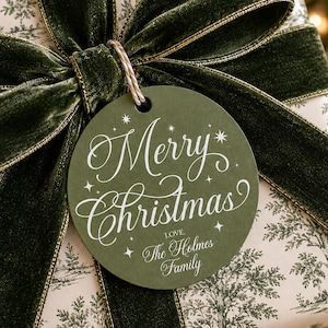 Peut inclure: Une étiquette cadeau ronde vert olive avec le texte blanc Merry Christmas et Love, The Holmes Family, attachée par une ficelle à un cadeau emballé dans du papier crème à motif d'arbres verts et noué avec un large ruban de velours vert foncé.