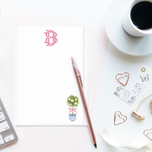 May include: A white notepad with a pink letter "B" monogram, a pink ribbon tied around a small lemon tree illustration, and a rose gold pen on a pink surface.